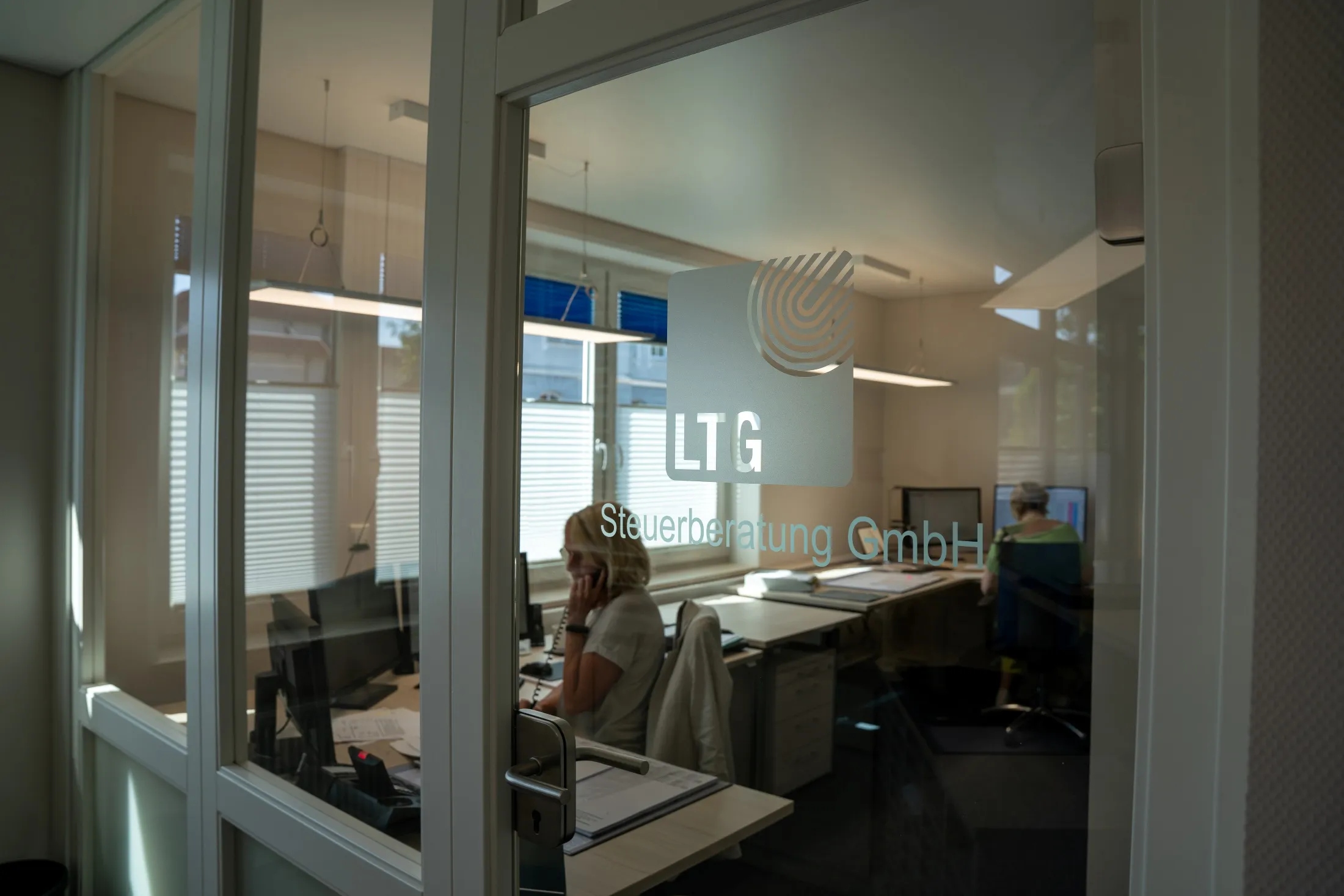 Glasbüro mit Besprechungstisch und Stühlen, eine Frau telefoniert
