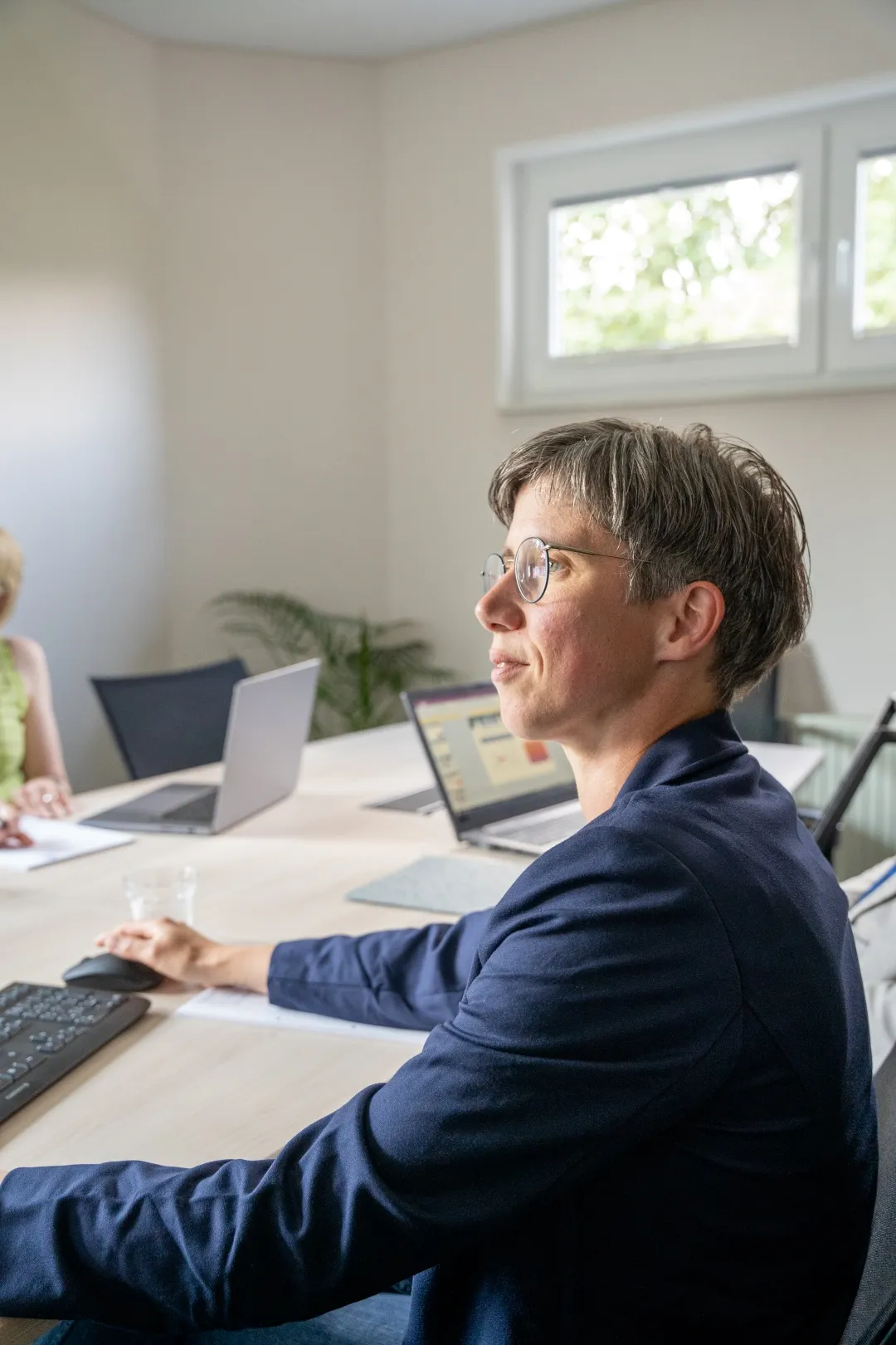 Fraumit kurzem Haar und Brille sitzt in einem Besprechungsraum an einem Tisch mit Laptop und Tastatur, schaut aufmerksam zur Seite.