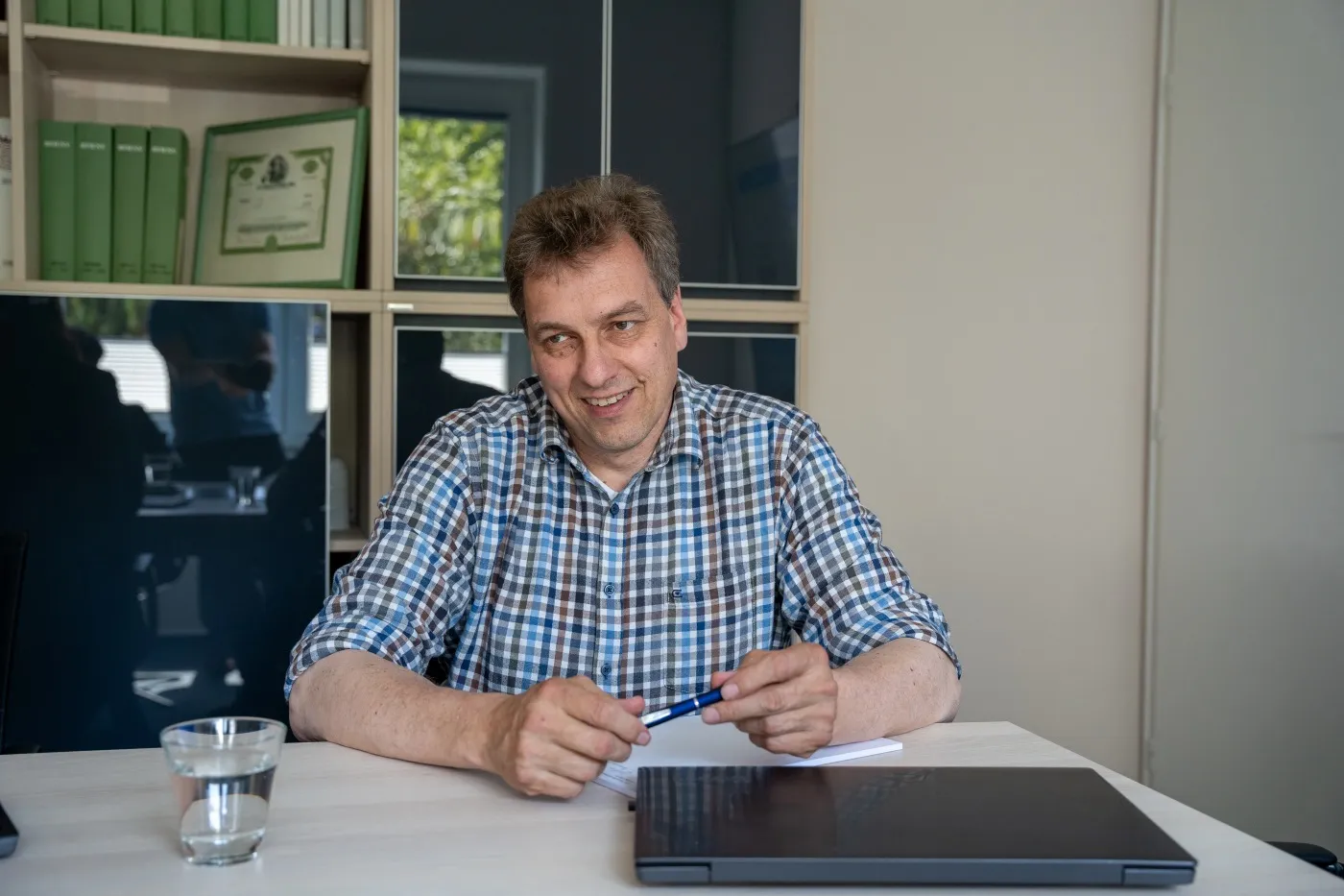 Mann mit kariertem Hemd sitzt an einem Tisch und lächelt
