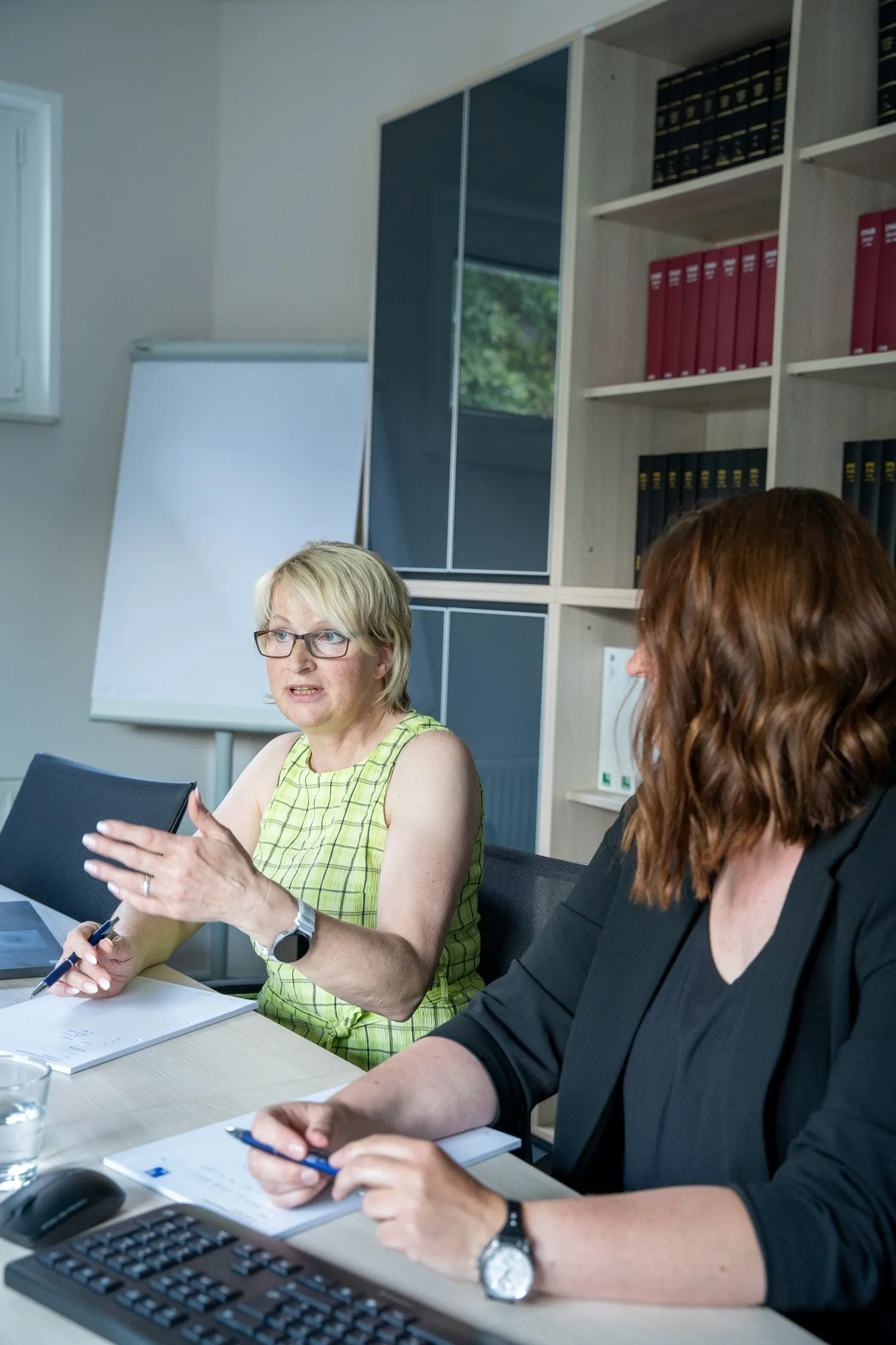 Zwei Frauen unterhalten sich an einem Tisch mit Aktenordnern im Hintergrund.
