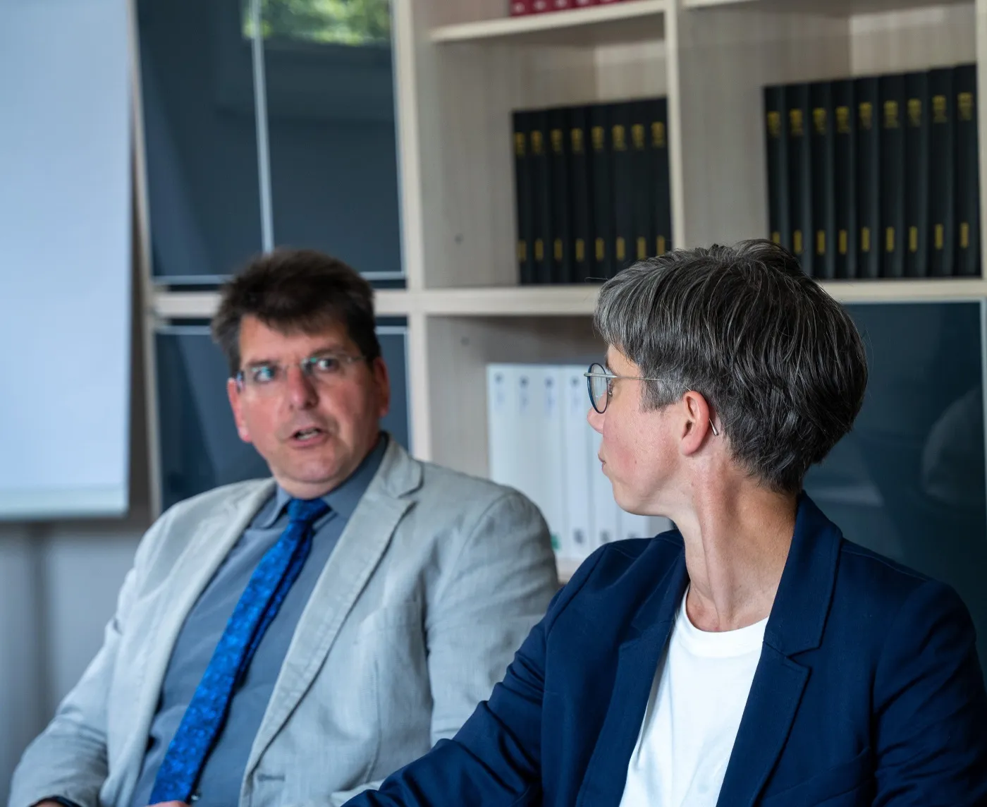 Zwei Personen im Gespräch in einem Büro mit Ordnern im Hintergrund
